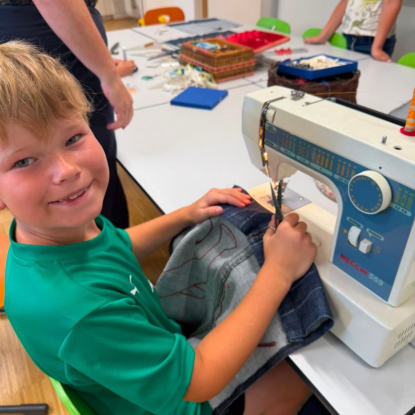A young boy is sitting at a sewing machine, smiling and holding scissors, while another person stands behind him.
