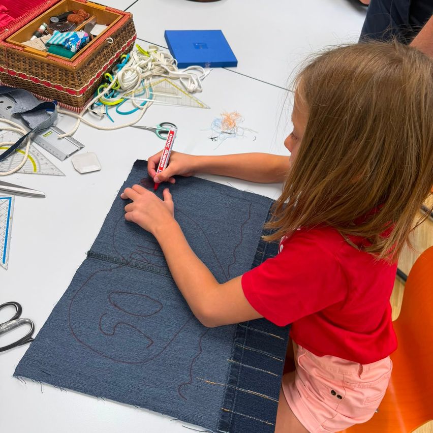 A young girl is sitting on a chair, drawing on a piece of denim fabric with a red marker. The table is cluttered with sewing tools and materials.