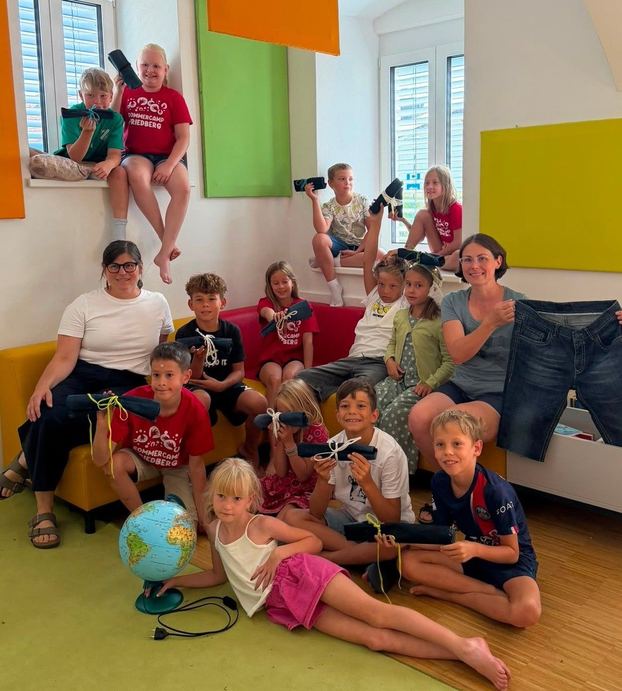 A group of children and adults are sitting and standing in a room, holding binoculars and a globe. Some are wearing glasses and sneakers.
