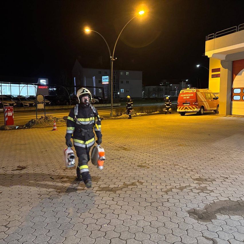 Ein Feuerwehrmann geht bei Nacht durch einen nassen Parkplatz und hält Ausrüstung in der Hand. In der Ferne gehen zwei weitere Feuerwehrleute auf einen Feuerwehrwagen zu.