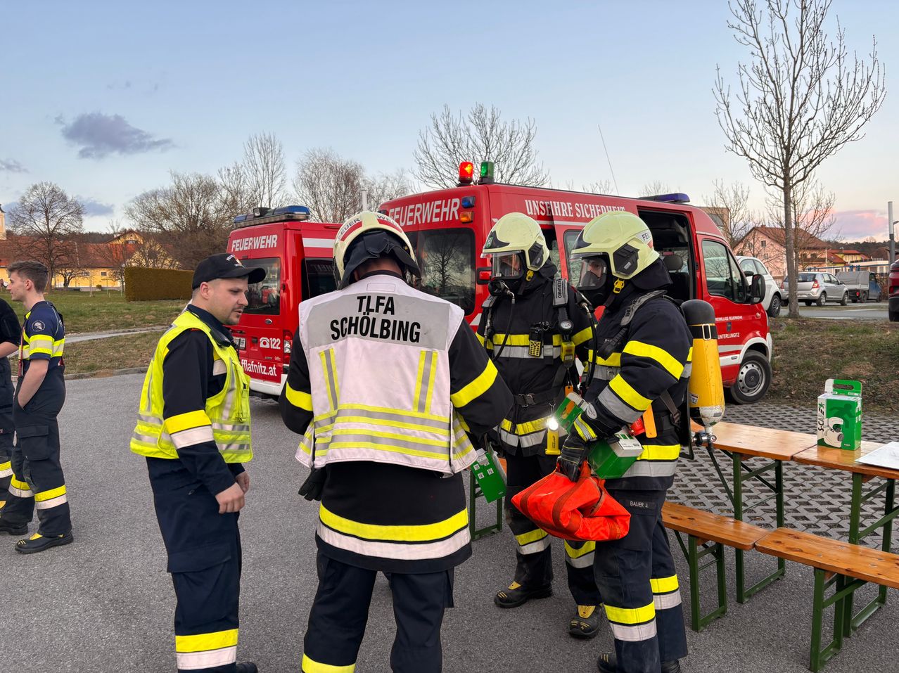 Feuerwehrleute in voller Ausrüstung stehen neben einem roten Feuerwehrauto. Einer hält eine Feuerlöscher, und zwei andere unterhalten sich mit einem Mann in einer reflektierenden Weste.