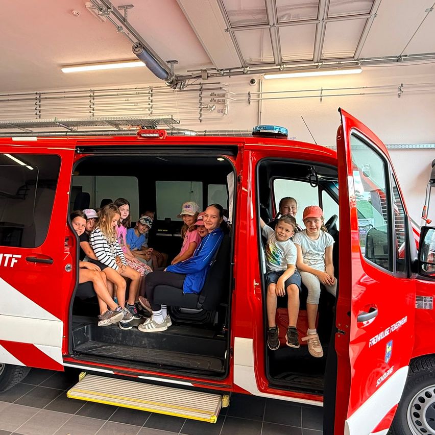 Eine Gruppe von Kindern sitzt in einem roten Feuerwehrauto, einige tragen Mützen und Turnschuhe, mit offenen Türen in einer Garage.
