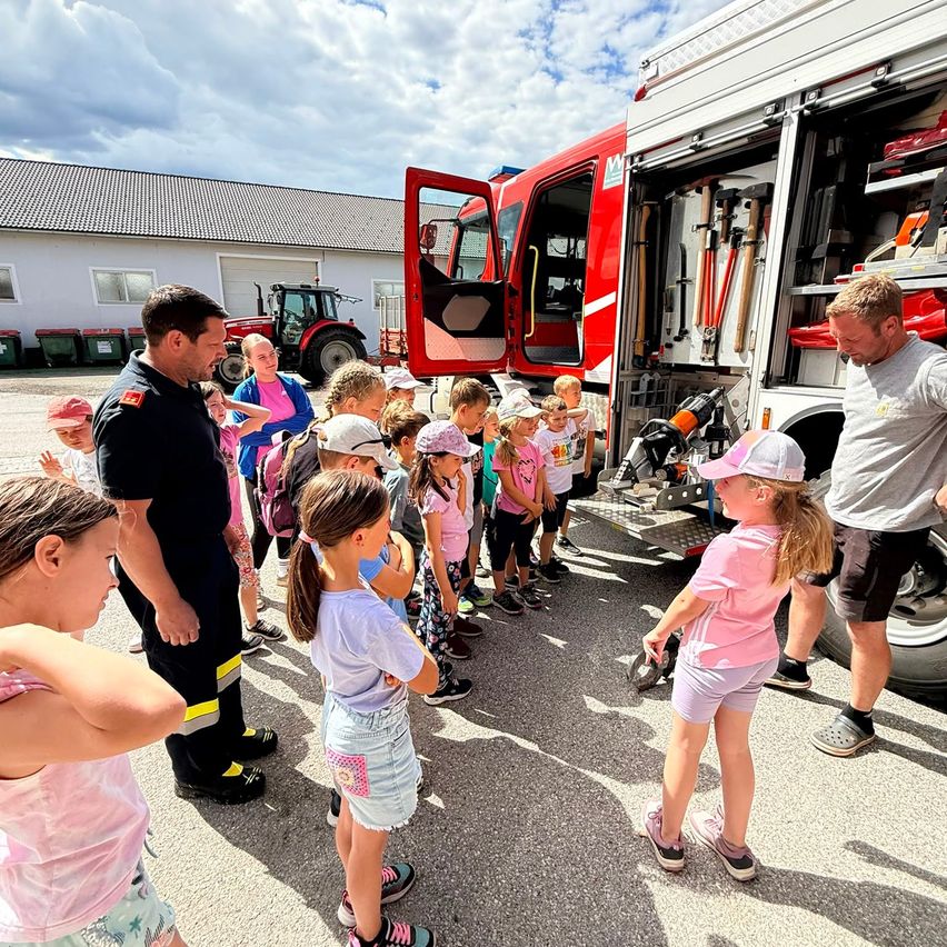 Eine Gruppe von Kindern und Erwachsenen versammelt sich um einen Feuerwehrwagen, wobei ein Feuerwehrmann ihnen etwas erklärt.