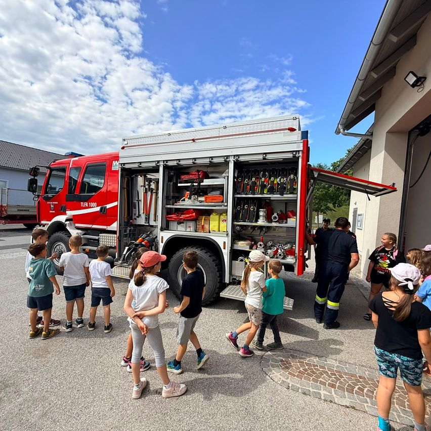 Eine Gruppe von Kindern und Erwachsenen versammelt sich um einen Feuerwehrwagen, dessen Tür offen ist und Ausrüstung im Inneren sichtbar ist. Die Kinder tragen Mützen und Turnschuhe, und die Erwachsenen stehen in der Nähe. Die Szene spielt sich im Freien ab, mit einem Gebäude und Bäumen im Hintergrund.