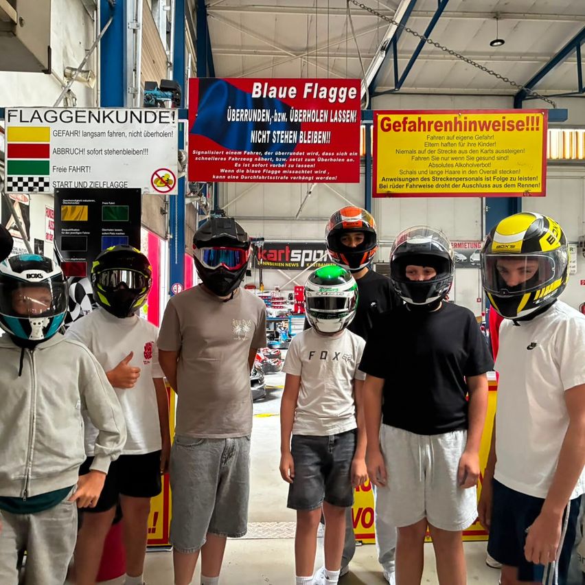Eine Gruppe junger Leute in Helmen und Schutzbrillen steht vor einer Kartbahn, mit Schildern, die Sicherheitsregeln und Anweisungen anzeigen.