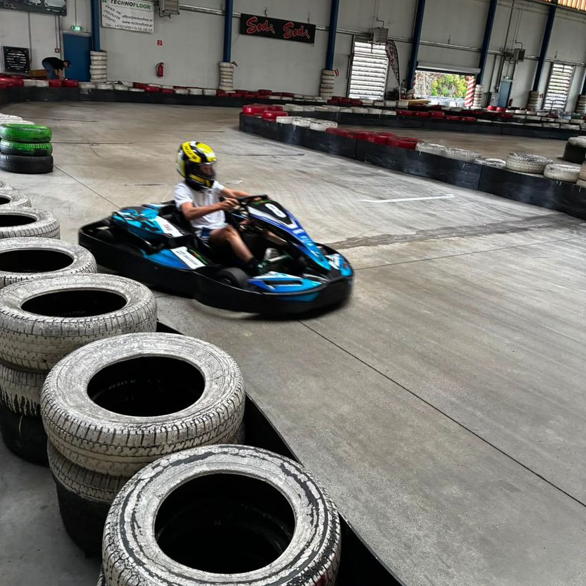 Eine Person mit einem gelben Helm fährt einen blauen Go-Kart auf einer Rennstrecke mit gestapelten Reifen an der Seite.