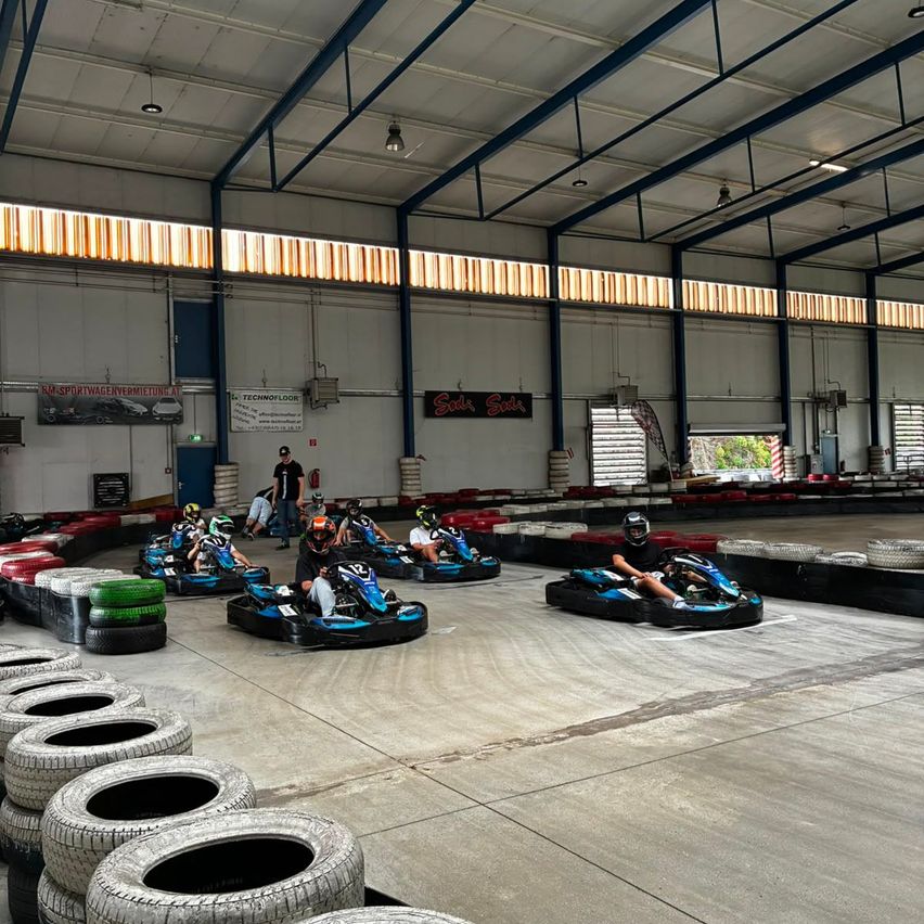 Mehrere Personen fahren Go-Karts in einer Indoor-Anlage. Sie tragen Helme und treten auf einer Rennstrecke an, die von Reifen umgeben ist. Die Anlage hat eine hohe Decke mit Lichtern und Bannern.