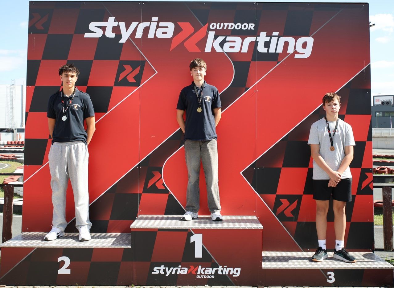 Drei junge Männer stehen auf einem Podium mit Medaillen um den Hals. Der mittlere Mann steht auf der zweiten Stufe, die anderen auf der ersten und dritten Stufe. Sie stehen vor einem roten und schwarzen Schachbretthintergrund mit dem Wort Styria Karting.