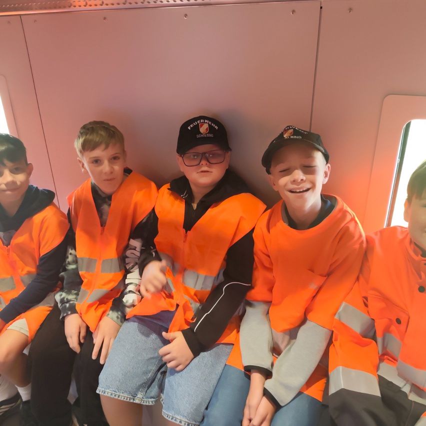 Fünf Jungen in orangefarbenen Sicherheitswesten und Mützen sitzen zusammen in einem Zug und lächeln für die Kamera.