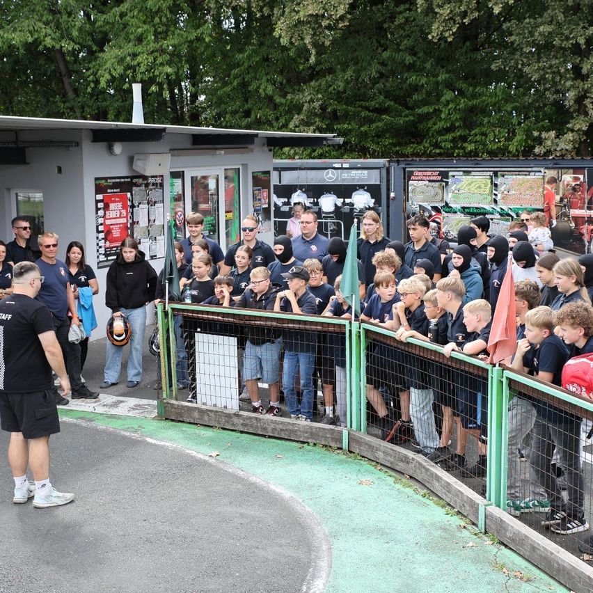 Eine Gruppe von Kindern und Erwachsenen beobachtet ein Rennen hinter einem grünen Zaun. Einige tragen Helme und Sonnenbrillen. Ein Mann in Schwarz steht vorne, möglicherweise gibt er Anweisungen.