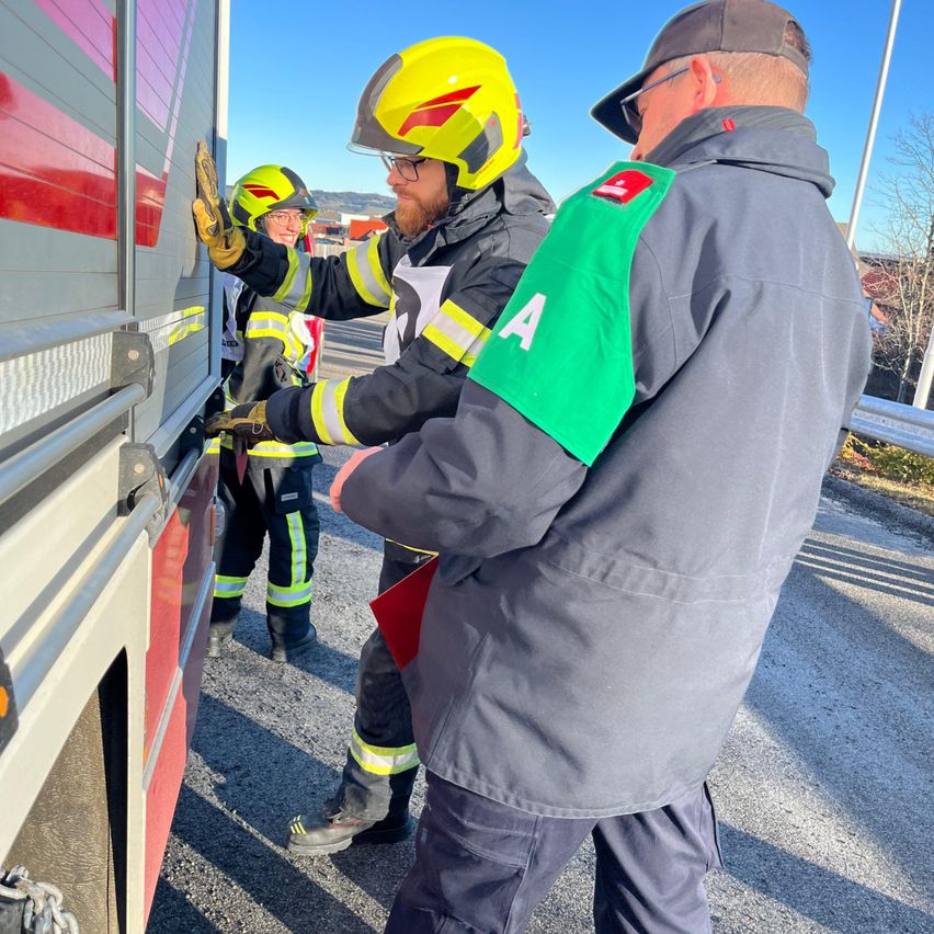 Zwei Feuerwehrleute in Schutzausrüstung untersuchen einen Feuerwehrwagen, einer hält die Tür offen und der andere hält eine Mappe.