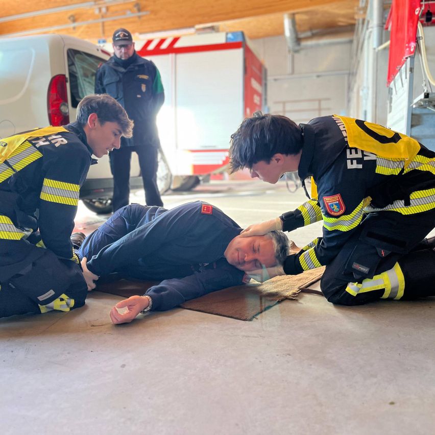 Zwei Feuerwehrleute in Uniform helfen einem Mann, der auf dem Boden liegt. Ein weiterer Feuerwehrmann steht hinter ihnen. Sie befinden sich in einer Garage mit einem weißen Van und einem Feuerwehrauto.