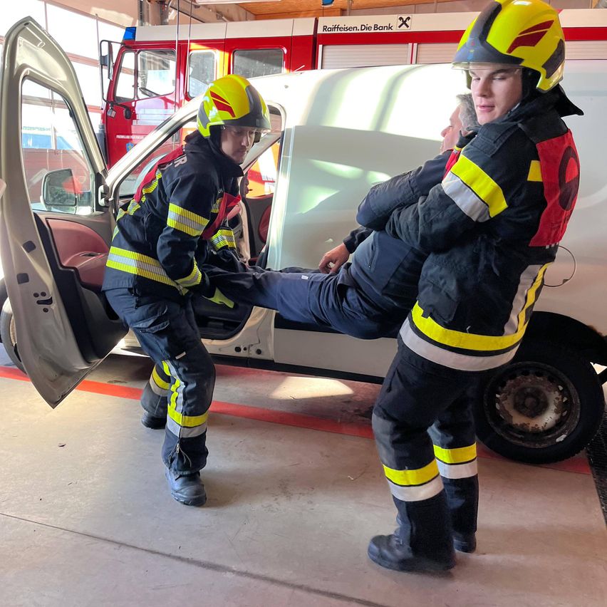 Zwei Feuerwehrleute helfen einem Mann, aus einem Fahrzeug in einer Garage auszusteigen, während ein Feuerwehrwagen dahinter geparkt ist.