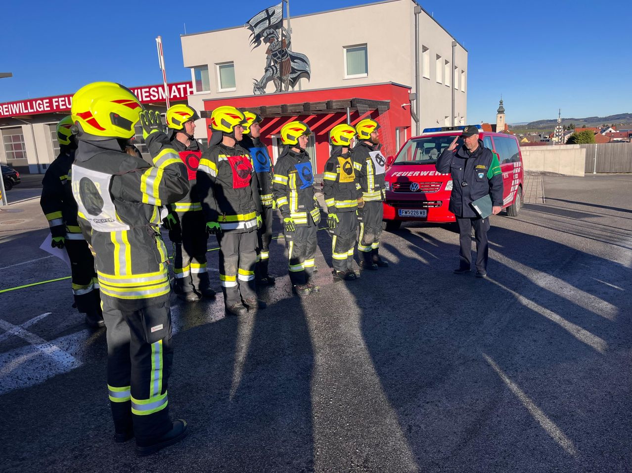 Eine Gruppe von Feuerwehrleuten steht vor einem Feuerwehrhaus mit einem roten Fahrzeug. Die Feuerwehrleute tragen gelbe Helme und Uniformen. Der Mann vorne hält eine Mappe und scheint eine Rede zu halten.