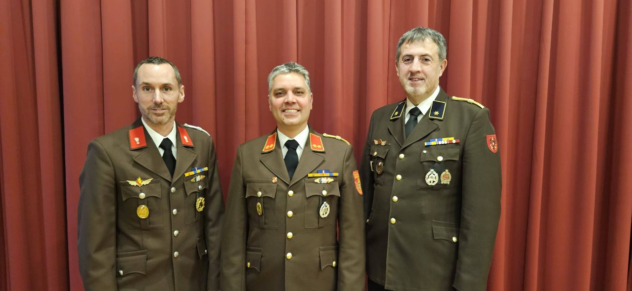 Drei Männer in Militäruniformen stehen nebeneinander und lächeln für ein Foto. Der Mann auf der linken Seite hat einen Schnurrbart und kurze Haare, während die anderen beiden längere Haare und Bärte haben. Sie tragen Medaillen und Abzeichen an ihren Uniformen.