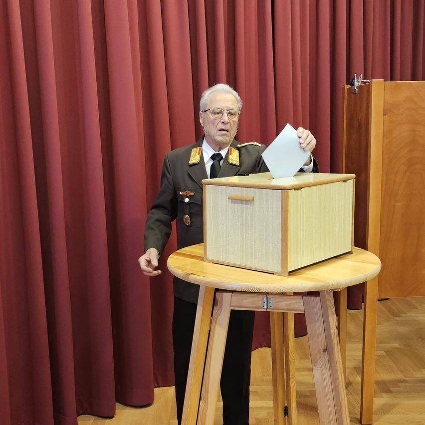 Ein älterer Mann in Uniform gibt seine Stimme in eine Holzurne auf einem Holztisch ab, mit roten Vorhängen im Hintergrund.