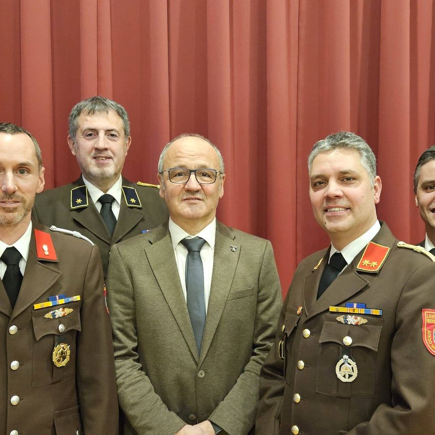 Fünf Männer in Uniformen stehen vor einem roten Vorhang und lächeln. Die zentrale Figur trägt eine Brille und eine Krawatte. Die Männer an den Seiten tragen Medaillen und Abzeichen.