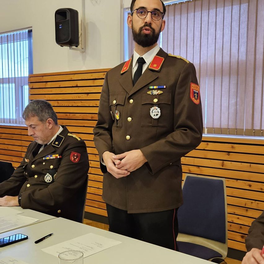 Ein uniformierter Offizier steht vor einem Tisch und sieht einem anderen Offizier gegenüber, der sitzt und Papiere betrachtet. Dahinter befindet sich eine Holzwand mit Fenster und Jalousien.