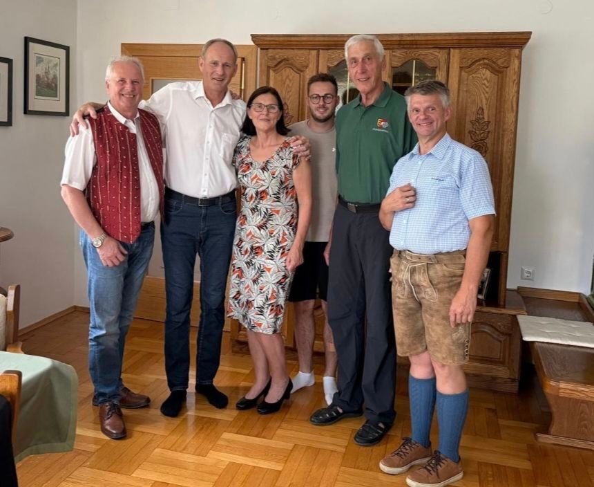 Sechs Erwachsene, darunter drei Männer und drei Frauen, stehen eng beieinander in einem Raum. Sie lächeln und scheinen für ein Foto zu posieren. Hinter ihnen steht ein hölzerner Schrank mit Glastüren.