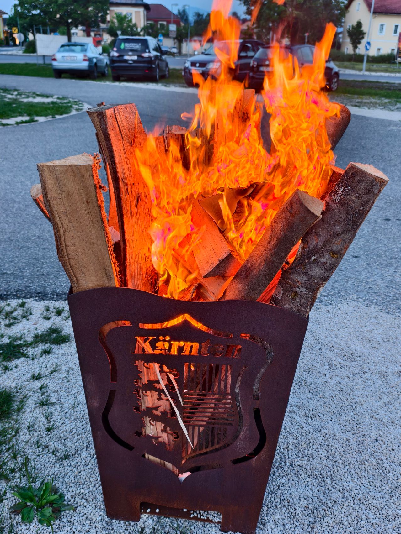 Ein Metallfeuerkorb mit brennendem Holz, der Name 'Kärnten' ist darauf. Dahinter sind Autos am Straßenrand geparkt.