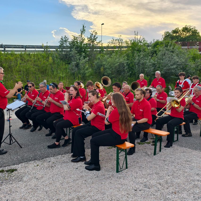 Ein Orchester von Musikern in roten Hemden und schwarzen Hosen spielt Instrumente in einer Freiluftumgebung, auf hölzernen Bänken sitzend und von einem Dirigenten begleitet.