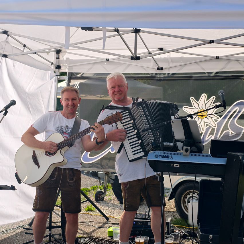 Zwei Männer stehen unter einem Zelt und spielen Gitarre und Akkordeon. Sie lächeln, mit einem Yamaha-Piano vor ihnen und einem Mikrofon. Im Hintergrund befindet sich ein Van.