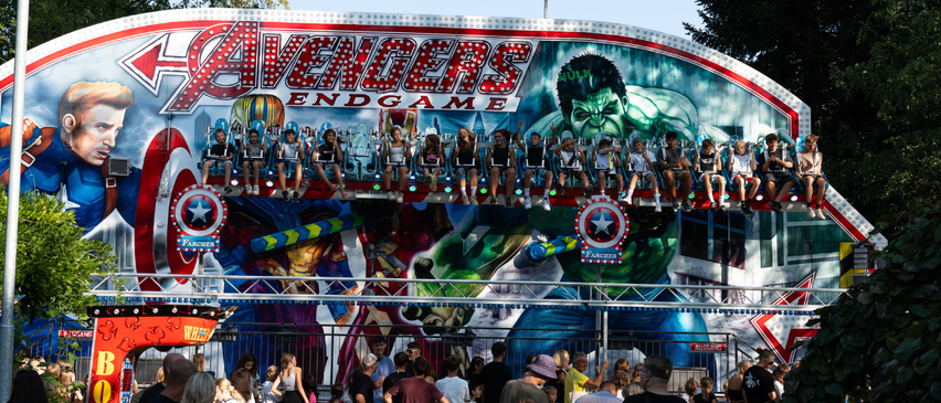 Eine Gruppe von Kindern fährt auf einer Avengers-inspirierten Achterbahn, wobei der Hulk prominent auf der Seite dargestellt ist. Viele Menschen beobachten die Fahrt.