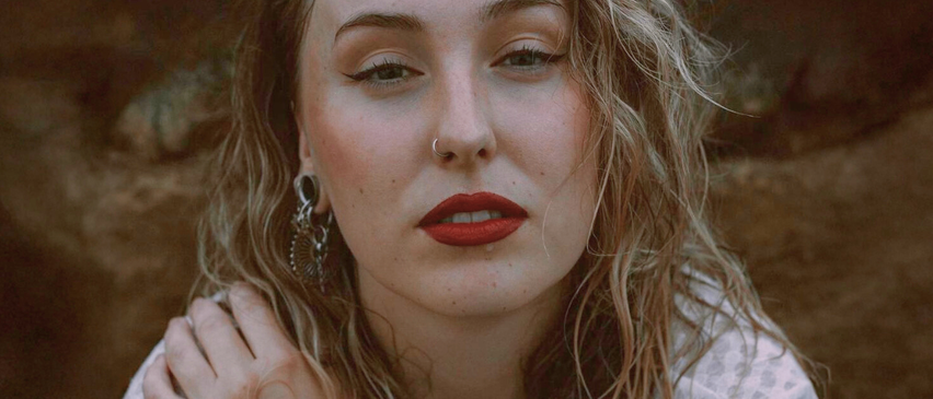 Ein Nahporträt einer Frau mit welligem Haar, rotem Lippenstift und einem silbernen Nasenring. Sie hat braune Augen und ein leichtes Lächeln.