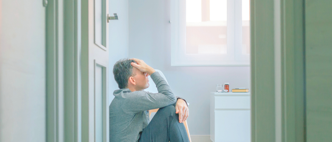 Ein Mann sitzt auf dem Boden mit dem Kopf in den Händen. Er trägt ein graues Sweatshirt und graue Hosen.