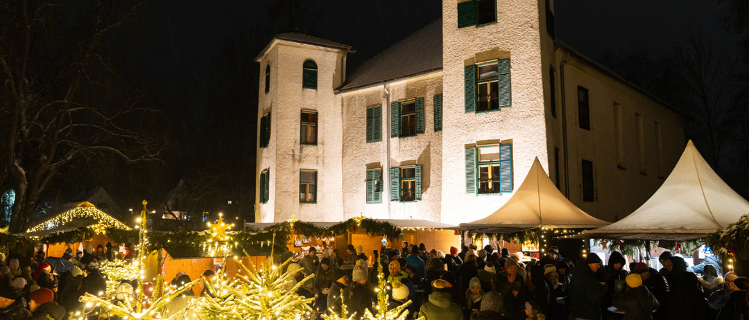 Ein Nachtmarkt unter einem großen Gebäude mit grünen Fensterläden und festlichen Dekorationen. Menschen versammeln sich um beleuchtete Weihnachtsbäume.