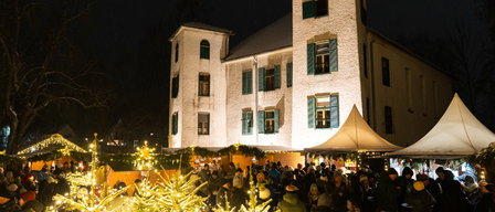 Ein Nachtmarkt unter einem großen Gebäude mit grünen Fensterläden und festlichen Dekorationen. Menschen versammeln sich um beleuchtete Weihnachtsbäume.