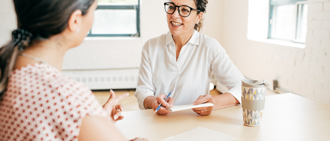 Eine Frau mit Brille sitzt an einem Schreibtisch, schreibt auf ein Blatt Papier, während eine andere Frau mit ihr spricht. Auf dem Schreibtisch befinden sich ein Stift, Papier und eine Kaffeetasse. Der Raum hat Fenster.