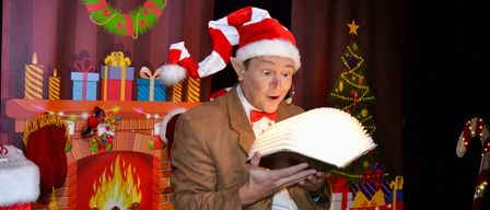 Ein Mann in Santa-Claus-Verkleidung liest ein leuchtendes Buch vor einem Kamin und einem Weihnachtsbaum, umgeben von Geschenken und Dekorationen.