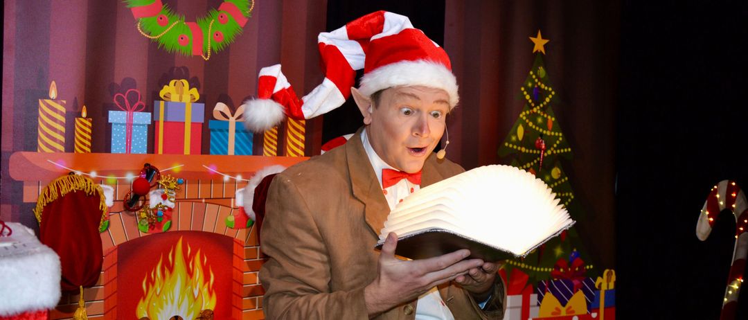 Ein Mann in Santa-Claus-Verkleidung liest ein leuchtendes Buch vor einem Kamin und einem Weihnachtsbaum, umgeben von Geschenken und Dekorationen.