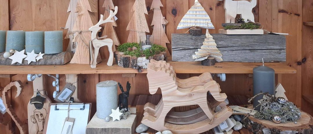 Ein Holzregal zeigt handgefertigte Weihnachtsdekorationen, darunter Bäume, Rehe und Kerzen. Über dem Regal hängen grüne Girlanden mit roten Kugeln. Holztiere und Bäume sind über die Regale verteilt.