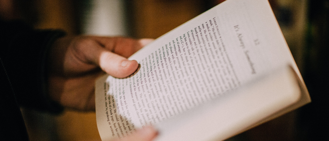 Eine Hand hält ein offenes Buch mit unscharfem Text, wobei ein bestimmter Abschnitt hervorgehoben wird. Der Hintergrund ist unscharf und betont das Buch.