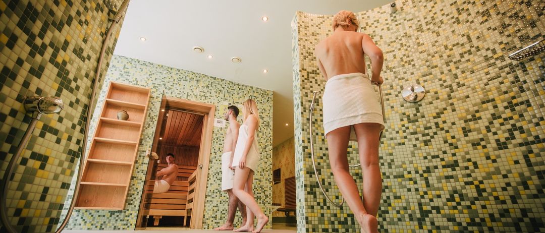 Drei Personen in einem Saunaraum, eine tritt ein, zwei sind dabei auszusteigen. Der Raum hat geflieste Wände und ein Holzregal.