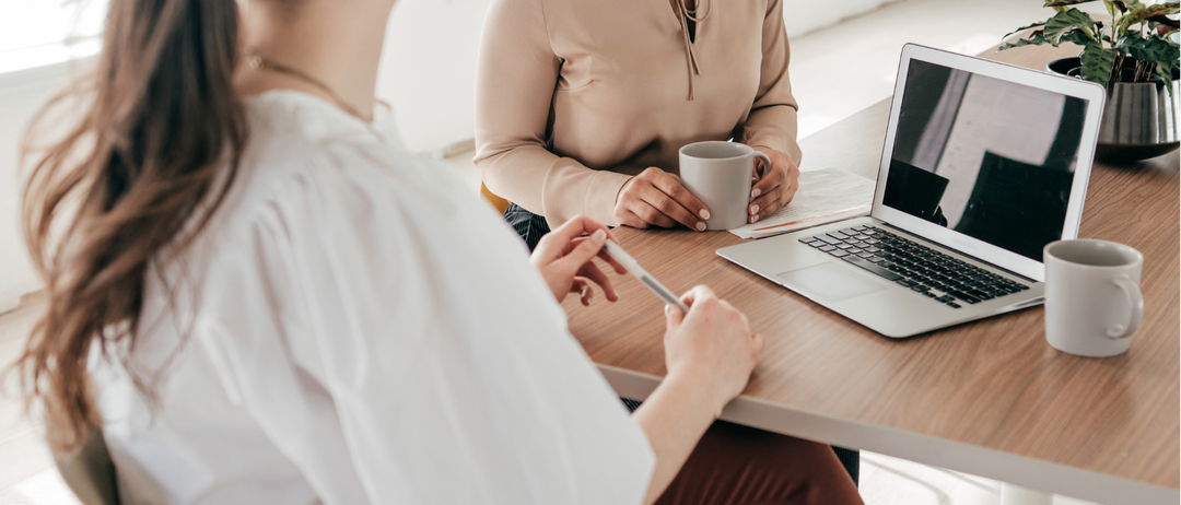 Zwei Frauen in Geschäftskleidung sitzen an einem Tisch mit einem Laptop, eine hält einen Stift und die andere eine Tasse. Die Frau auf der rechten Seite schaut auf den Laptop.