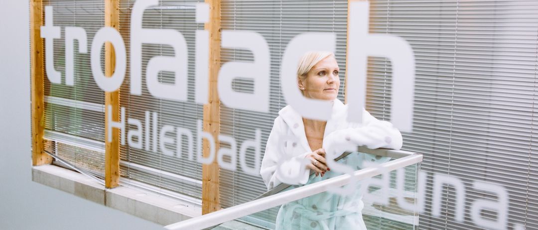 Eine Frau in einem weißen Bademantel mit Punkten steht auf einem Balkon, lächelt und hat die Hände in der Taille. Hinter ihr befindet sich ein Gebäude mit einem Schild, das 'Hallenbad' steht.