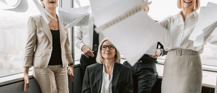 Mehrere Personen in formeller Kleidung in einem Raum, eine Frau lacht, während eine andere Papiere in die Luft wirft.