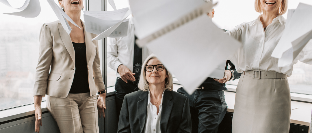 Eine Gruppe von Menschen steht in einem Büro, eine Frau lacht und hält Papiere. Eine andere Frau mit Brille sitzt mit ausgestreckten Armen. Papiere fliegen in der Luft.