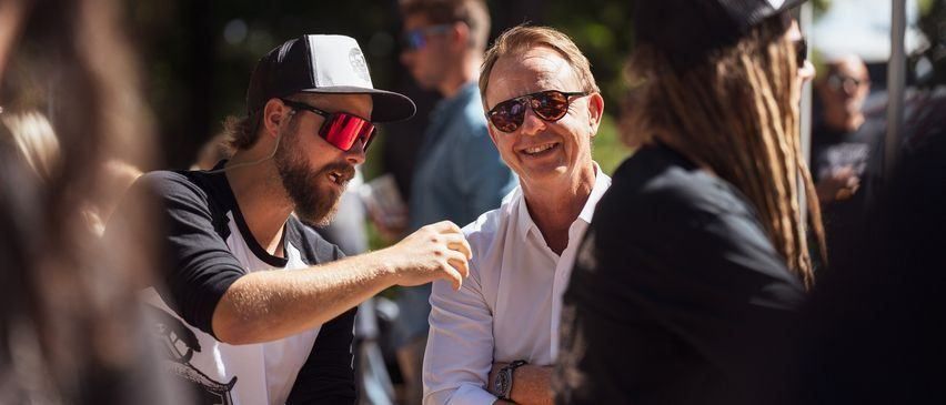 Zwei Männer mit Bärten, einer in einem weißen Hemd und der andere in einem schwarzen Hemd, lächeln und unterhalten sich miteinander.