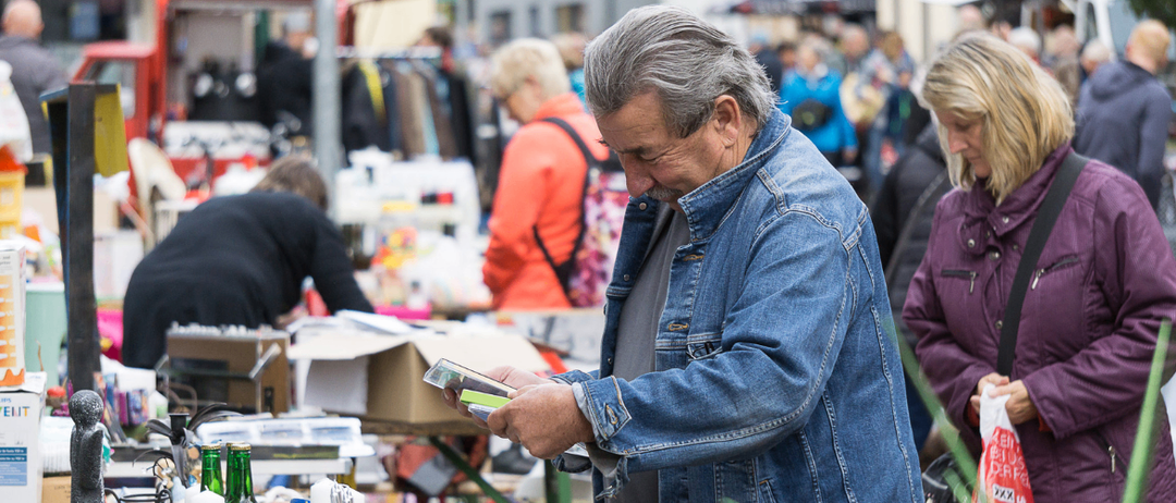 Ein älterer Mann betrachtet einen Gegenstand auf einem Flohmarkt, mit verschwommenen Menschen und Ständen im Hintergrund.