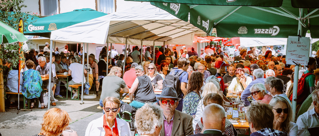 Eine große Menschenmenge versammelt sich unter grünen Sonnenschirmen in einem Biergarten, gefüllt mit Tischen und gesprächigen Menschen. Verschiedene Getränke stehen auf den Tischen.