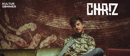 Ein Mann mit Bart und Schnurrbart sitzt auf einem Sofa mit einer Gitarre. Er trägt ein Camouflage-Hemd und Jeans. Er schaut nach rechts. Hinter ihm befindet sich ein verschwommenes Bild einer Wand mit Wellenmuster.