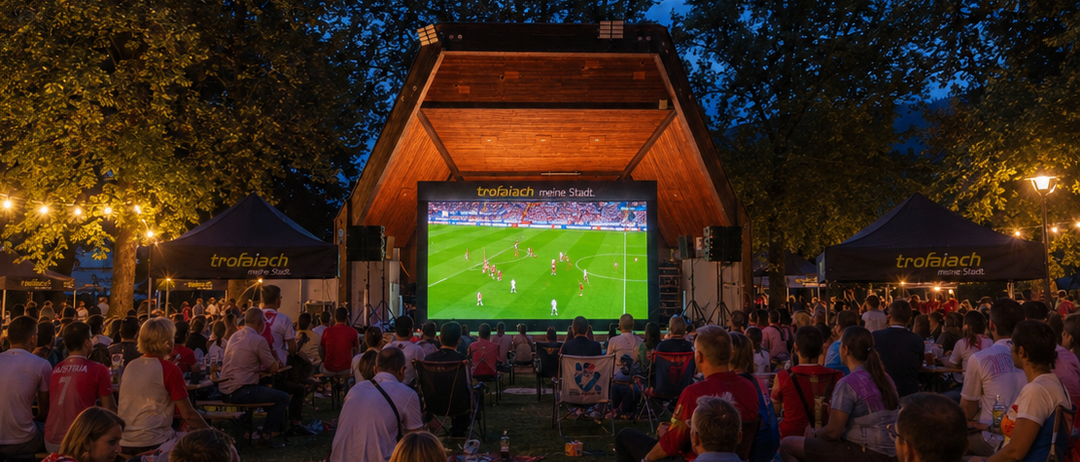 Eine Menschenmenge schaut ein Fußballspiel auf einer großen Leinwand draußen bei Nacht. Die Leute sitzen auf Stühlen und Decken, und es gibt Bäume und Zelte im Hintergrund.