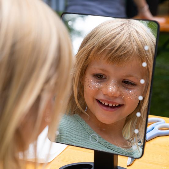 Ein kleines Mädchen mit blonden Haaren und Glitzer auf dem Gesicht lächelt in einen Spiegel. Sie trägt ein grünes Shirt. Hinter ihr ist eine verschwommene Person zu sehen.