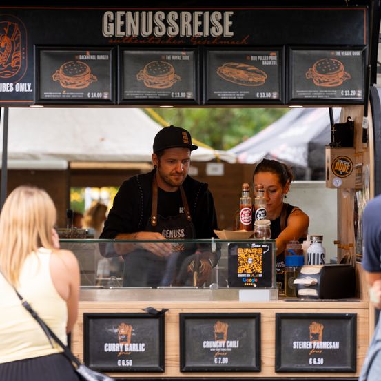Ein Burger-Truck namens Genussreise. Ein Mann in einer schwarzen Schürze bereitet hinter einer Glasscheibe Essen zu. Eine Frau sitzt an der Theke und eine blonde Frau schaut sich das Menü an. Verschiedene Burger-Optionen sind auf der Menütafel angezeigt.