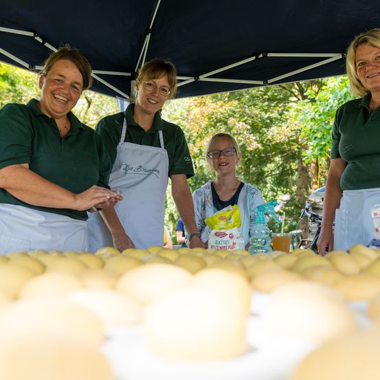 Drei Frauen in grünen Uniformen und Schürzen stehen unter einem Zelt. Sie lächeln. Eines Mädchen in der Mitte hält eine Tüte. Es gibt viele gelbe Bälle auf dem Tisch vor ihnen.