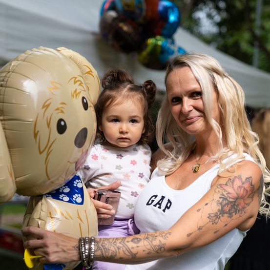 Eine Frau hält einen großen, hundförmigen Ballon und steht mit einem kleinen Mädchen. Das Mädchen hält einen kleinen Becher. Hinter ihnen steht ein weißes Zelt mit bunten Luftballons.
